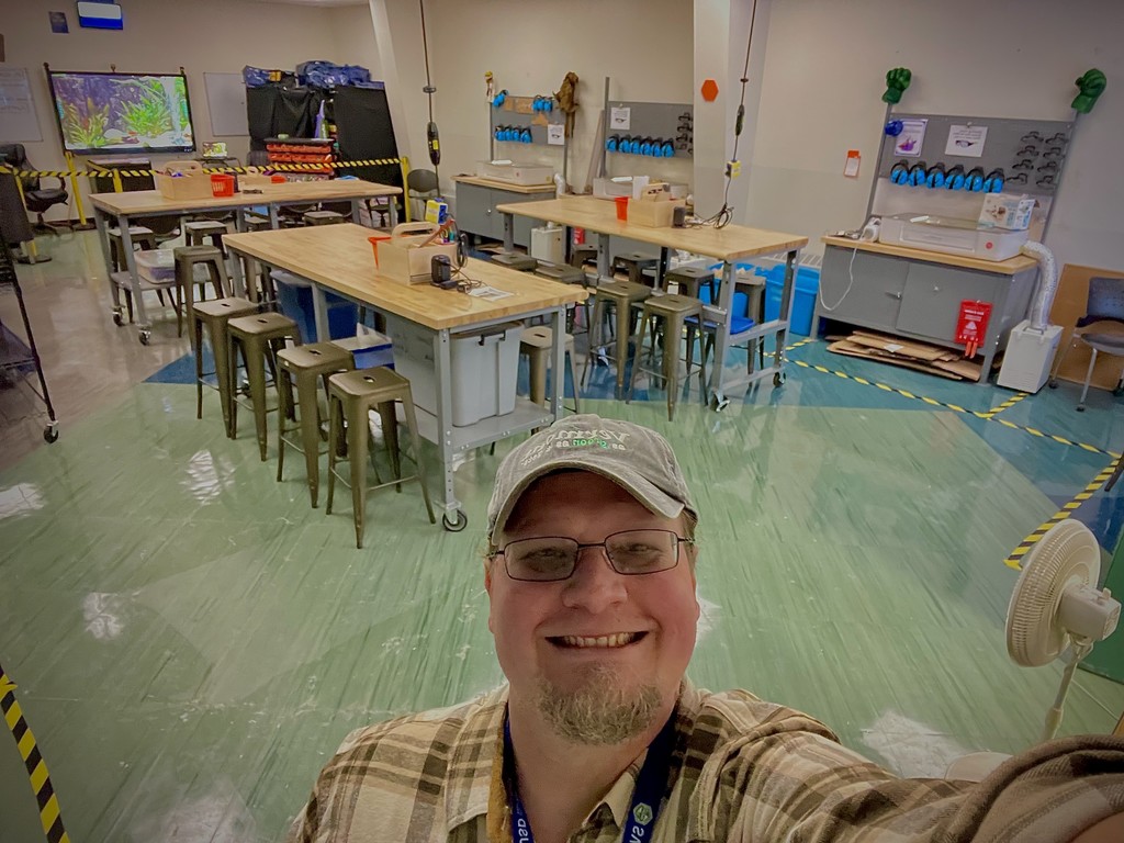 A smiling teacher takes a selfie in a clean and organized classroom with tall tables, stools, and workstations.