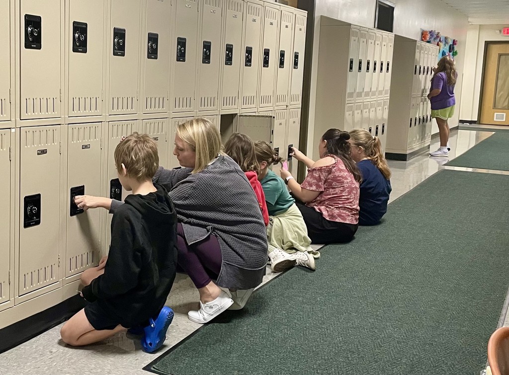 Several students kneel at lockers practicing combinations, with two adults assisting them in the hallway.