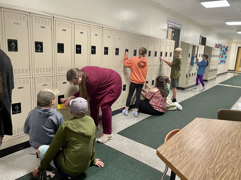A teacher helps two students open their lockers while other children practice on nearby lockers in a school hallway.