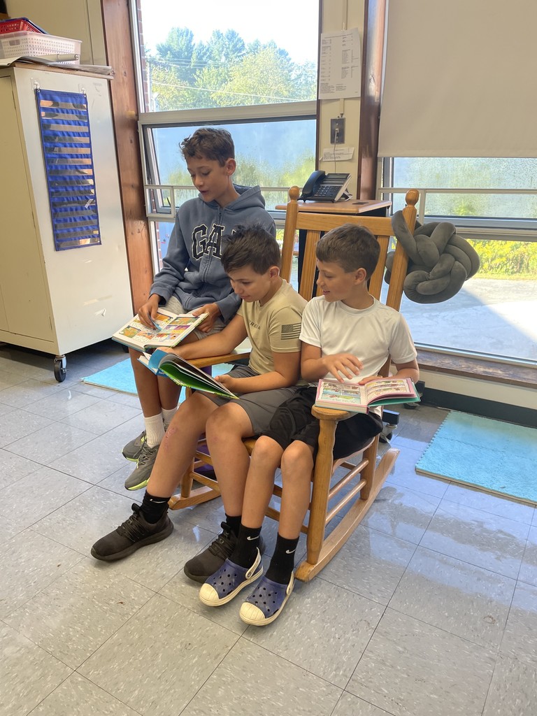 Three boys sit together in a wooden rocking chair, reading books in a classroom.