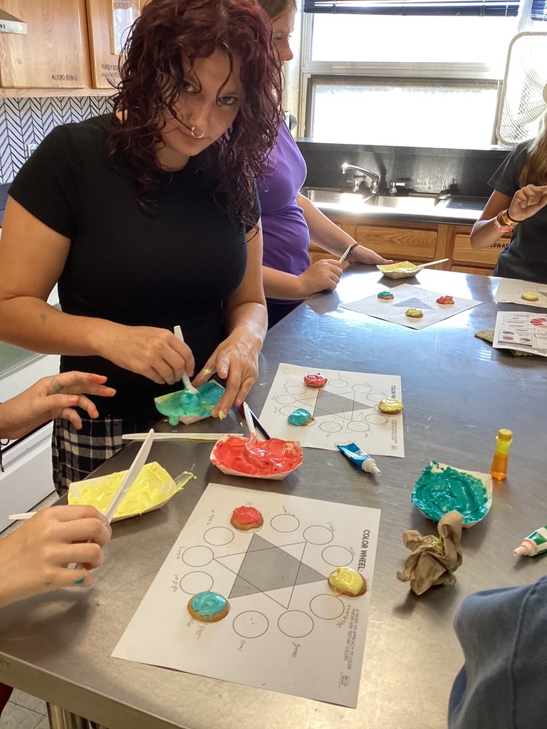 A student spreads brightly colored frosting on cookies while using a color wheel activity sheet in a classroom kitchen.