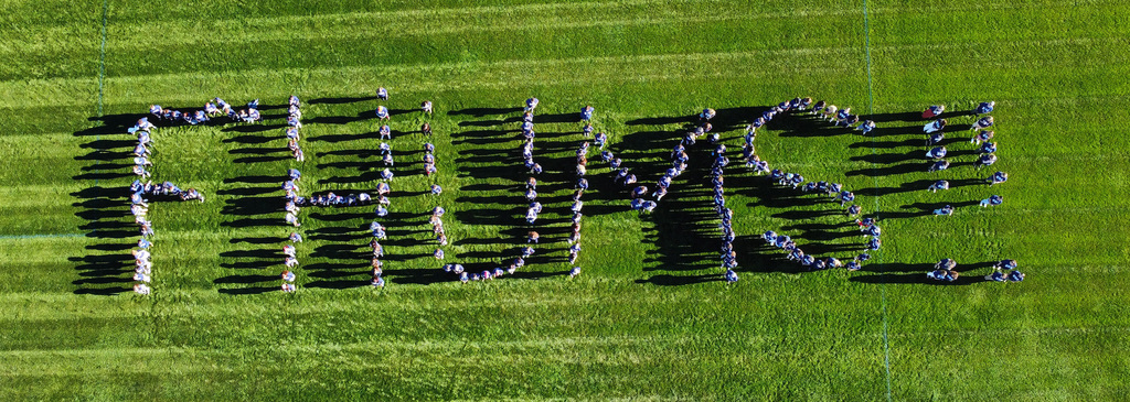 Aerial view of students standing on a grassy field arranged to spell out FHUMS.