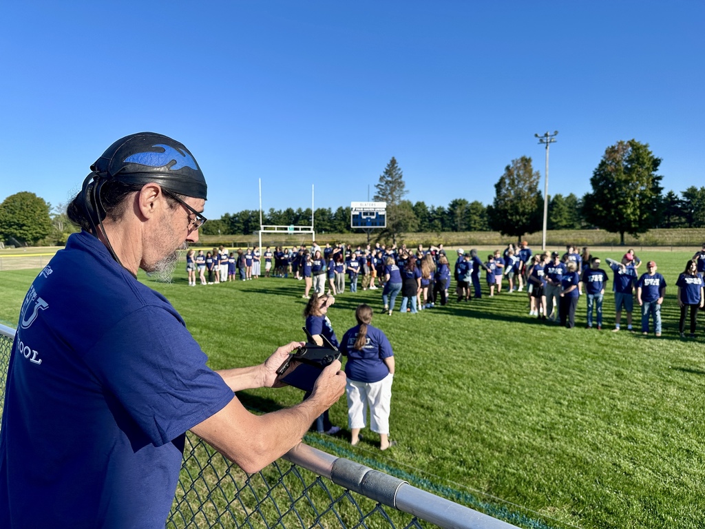 A man operates a Drone controller while students and staff gather on the field in FH shirts.