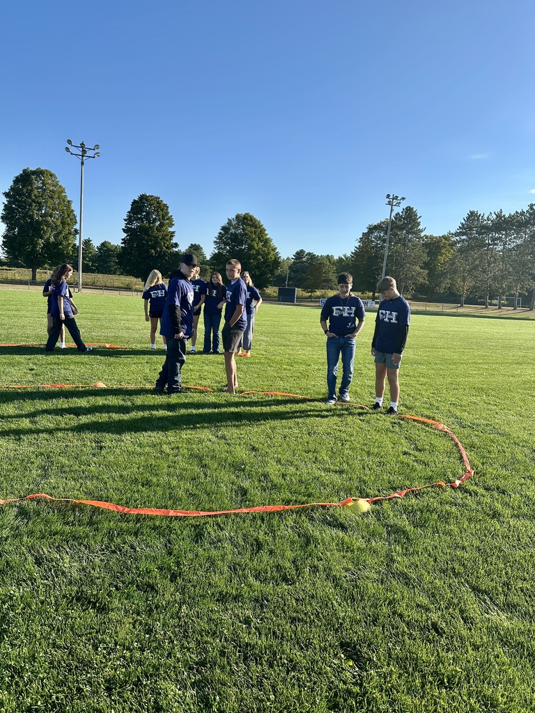 A group of students wearing matching FH shirts stands inside a large circle outlined on the grass.