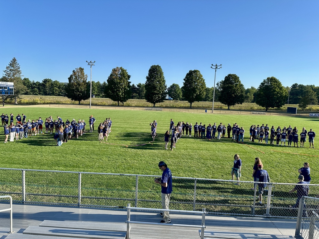 Groups of students and staff in FH shirts begin forming shapes on the field, viewed from the bleachers.