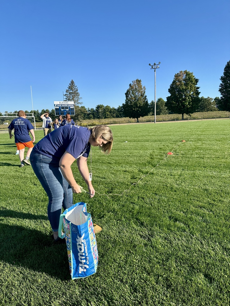 A woman bends down on a football field setting up string and markers, with others preparing in the background.