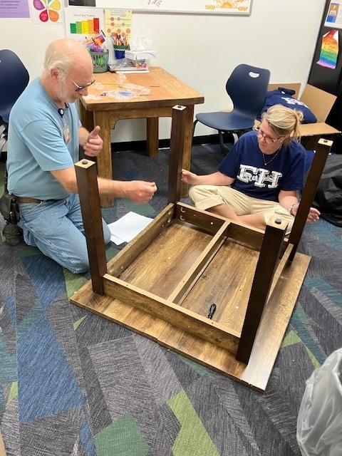 Two adults kneel on the floor assembling a wooden table upside down in an office