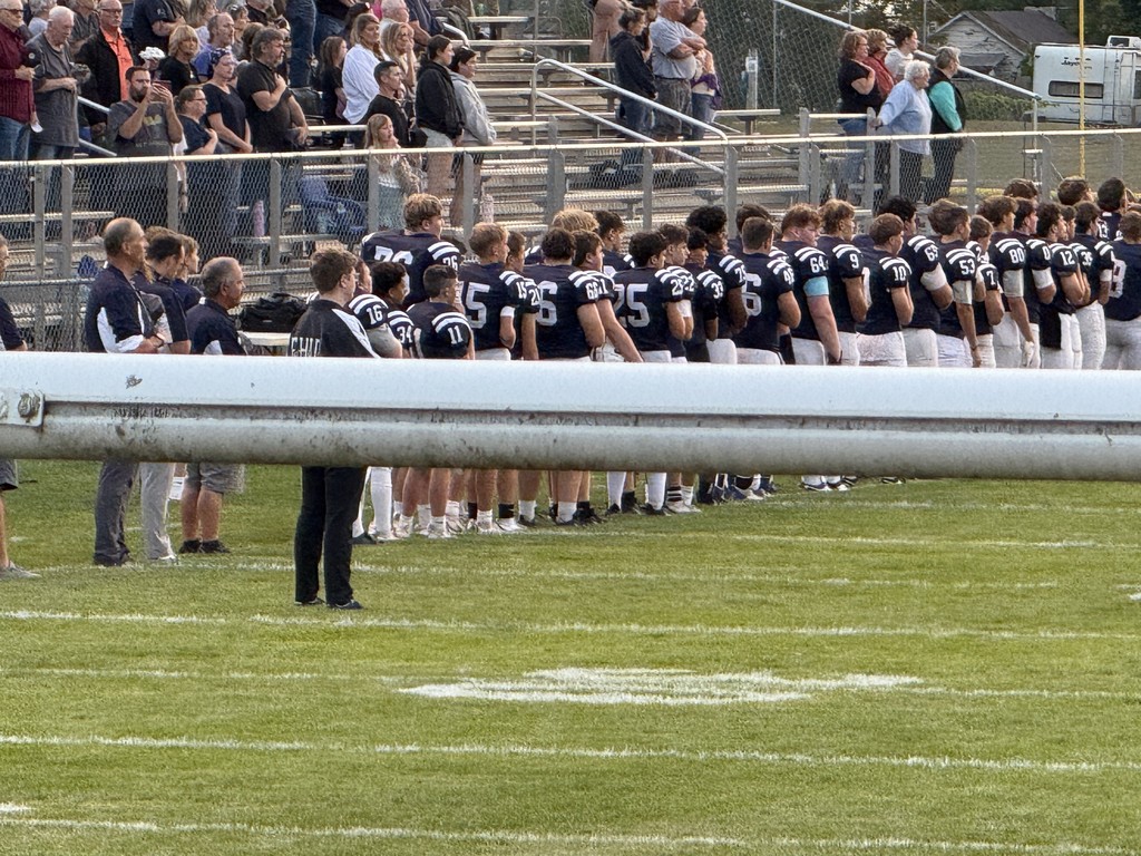 Fair Haven Union football players and coaches line up on the field with fans standing in the bleachers during the national anthem.