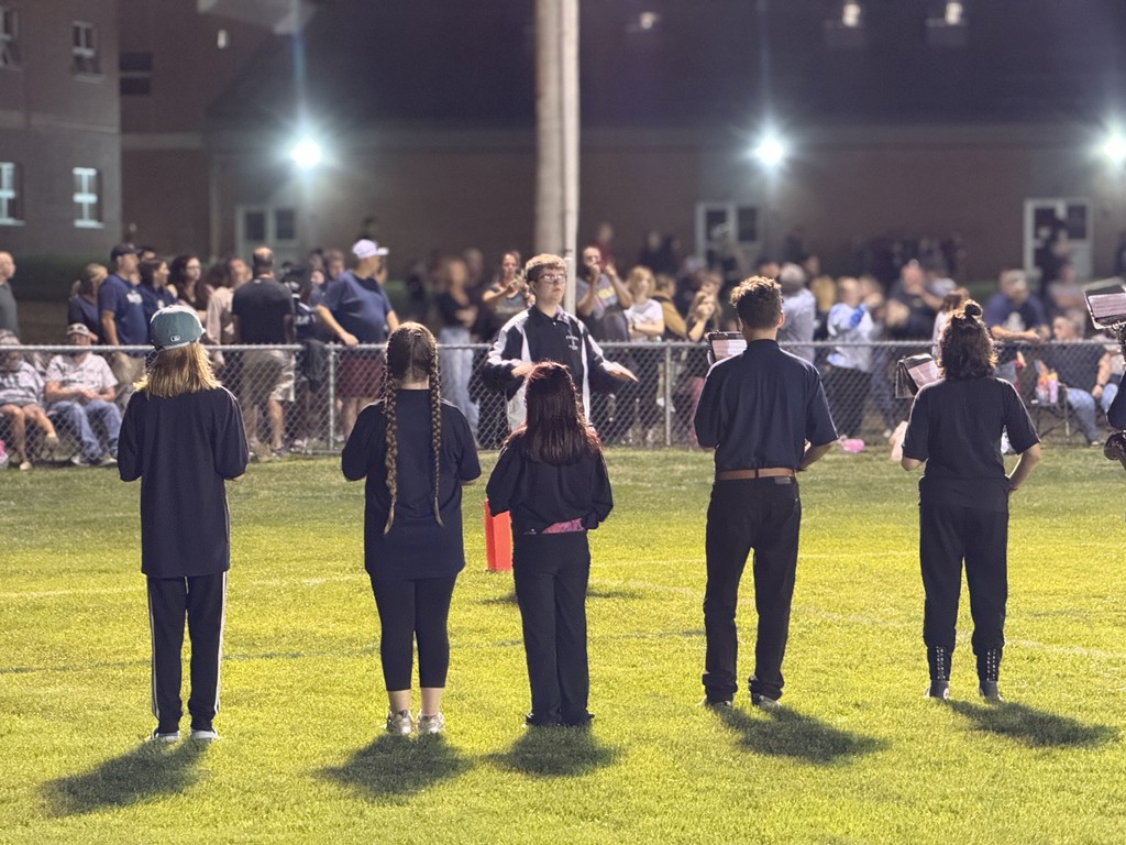 Student conductor directs the band on the field as musicians play in front of a cheering crowd.
