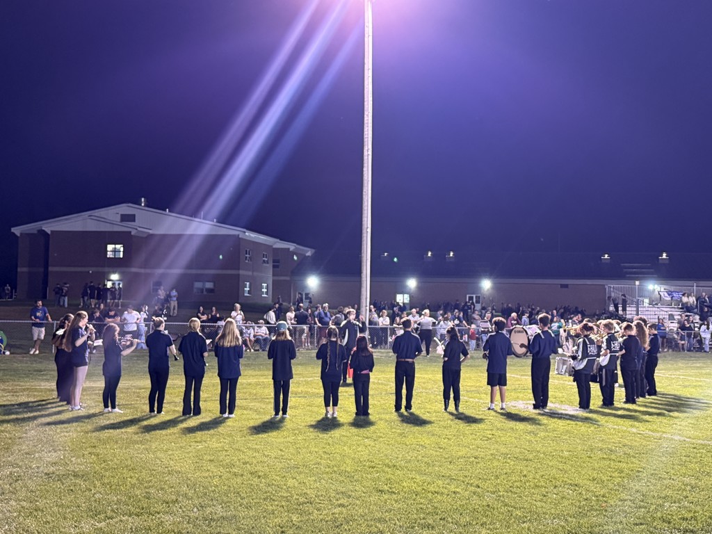 Nighttime view of the marching band performing with the audience and school building in the background.