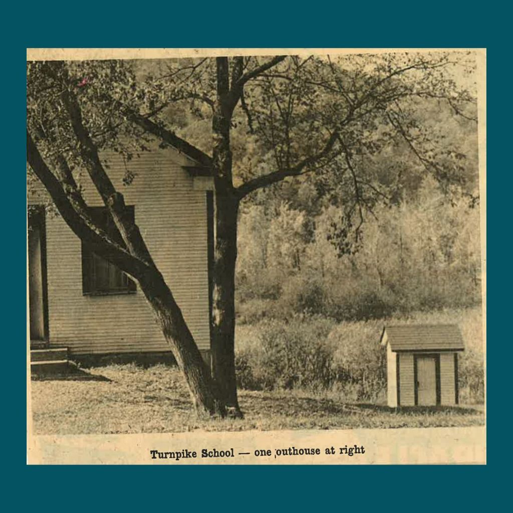 Black and white photo of Turnpike School with a small outhouse to the right, surrounded by trees and grass.