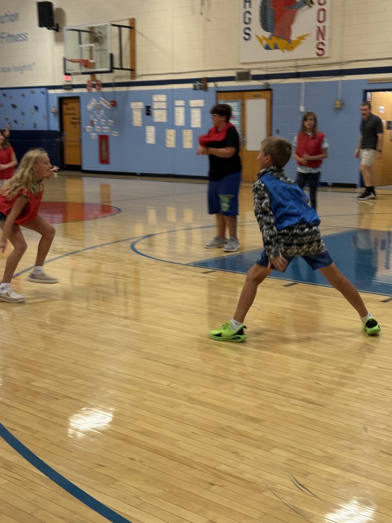 Students wearing pinnies play a game called fill the castle in a school gymnasium, with a hoop and equipment on the floor nearby. Students need to capture the equipment in the hoop without being tagged.