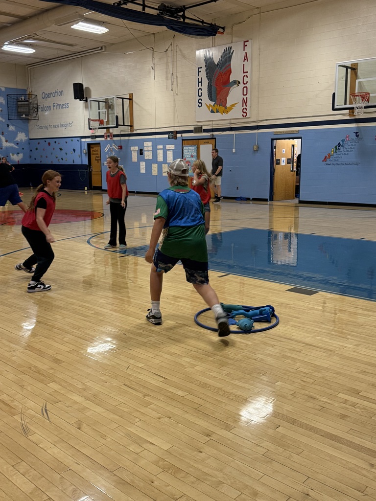 Students wearing pinnies play a game called fill the castle in a school gymnasium, with a hoop and equipment on the floor nearby. Students need to capture the equipment in the hoop without being tagged.