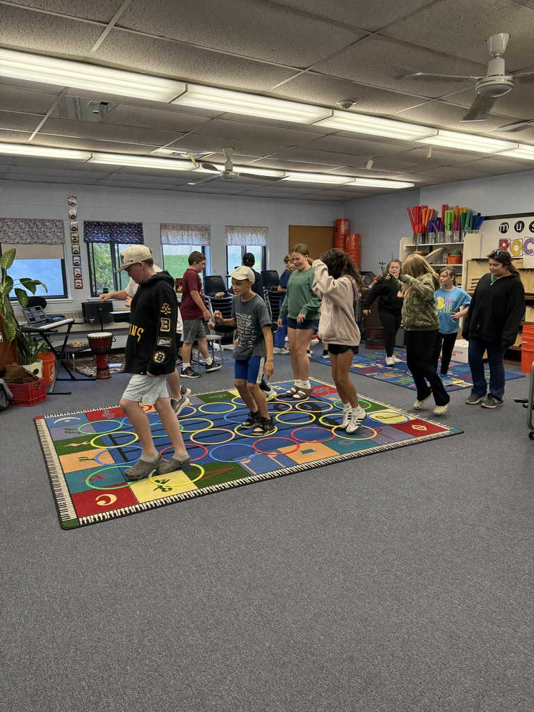 Students stand and move together, based on the beat and rhythm of the music playing, on a colorful alphabet rug in a classroom.