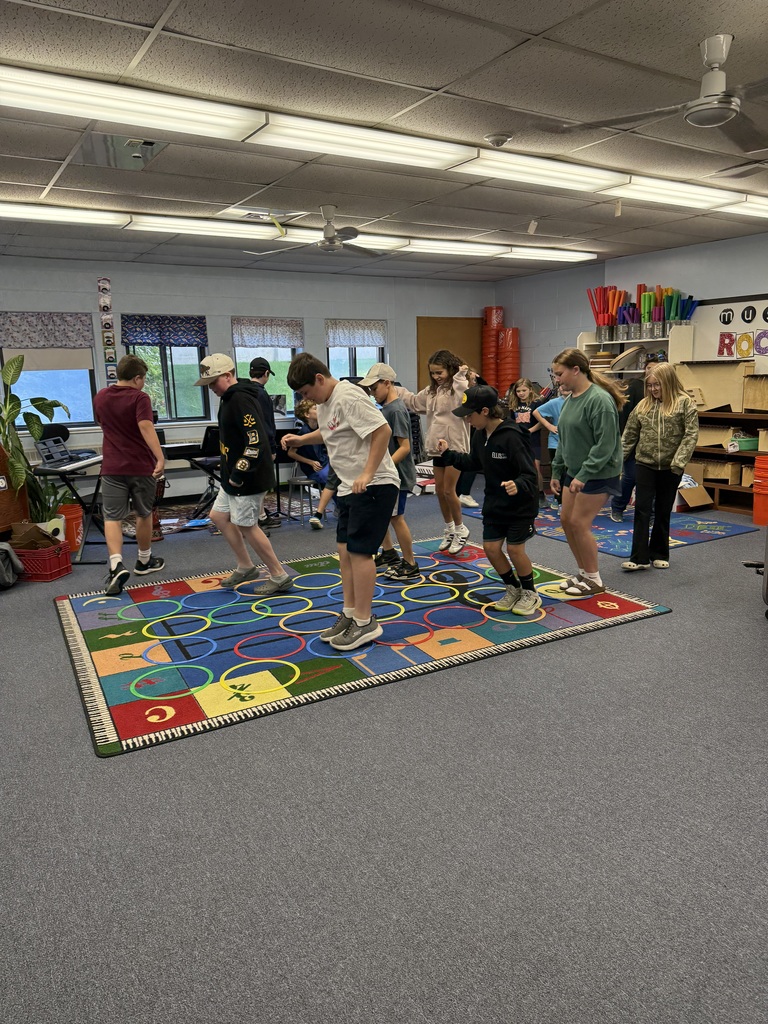 Students stand and move together, based on the beat and rhythm of the music playing, on a colorful alphabet rug in a classroom.