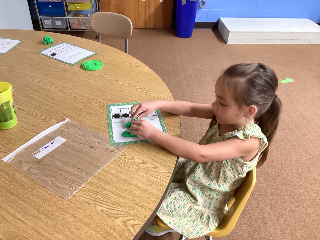 A young girl sits at a table using green Play-Doh on a counting mat.