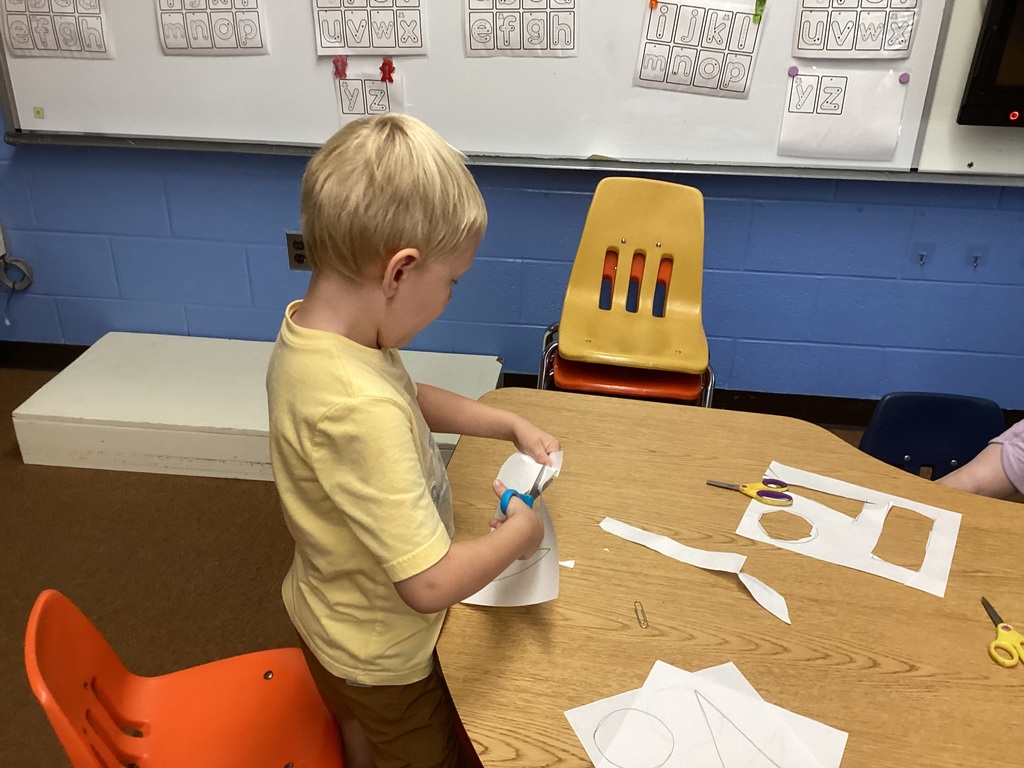 A young boy in a yellow shirt cuts out shapes from paper with scissors at a classroom table.