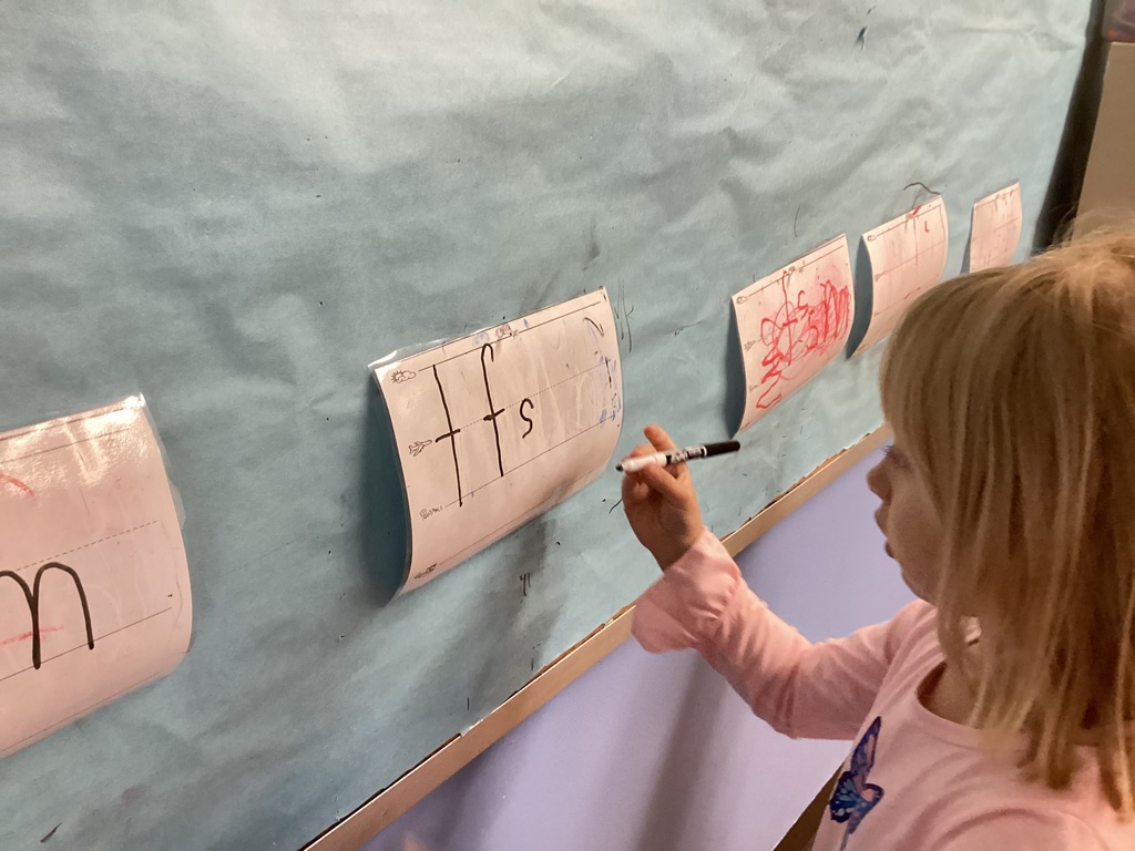 A student uses a marker to trace letters on a worksheet displayed on a bulletin board.