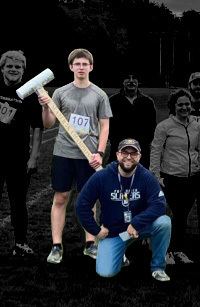 Two people at the Slater 5K: one standing and holding a large mallet, the other kneeling in front wearing a Slaters sweatshirt and cap. Background is faded with other participants visible.