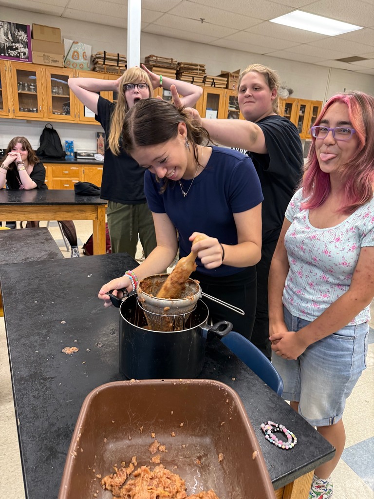 A group of students smile and laugh while straining applesauce; one student sticks out their tongue playfully.