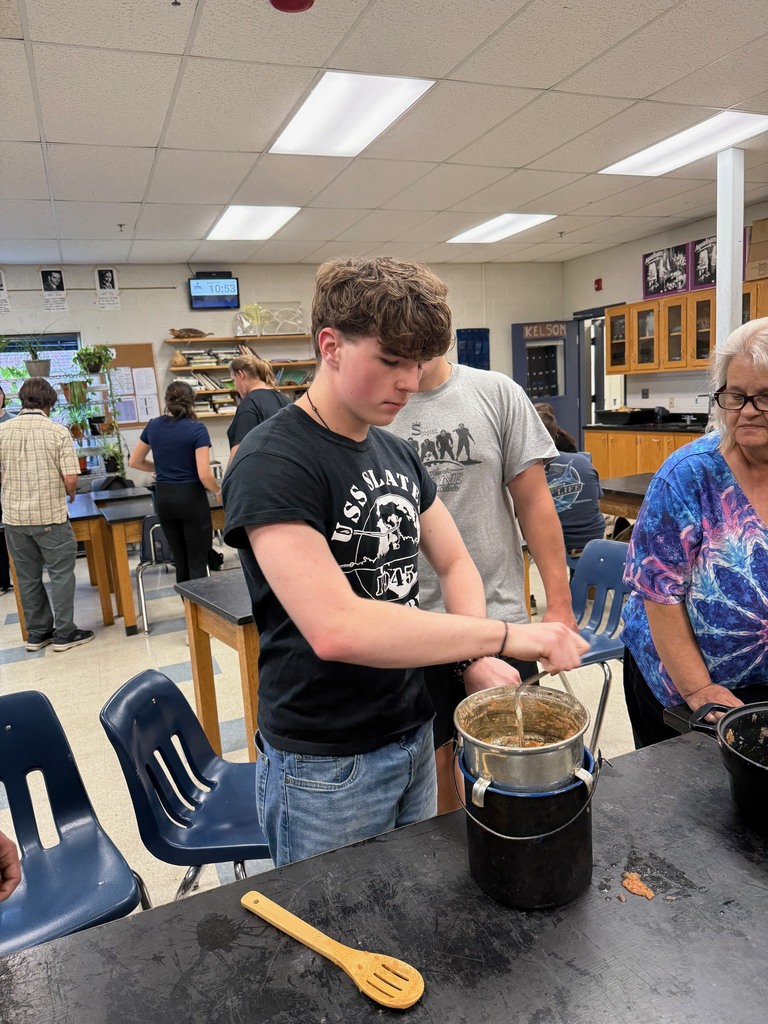 A student uses a strainer to press apples into a pot with classmates and teacher nearby.