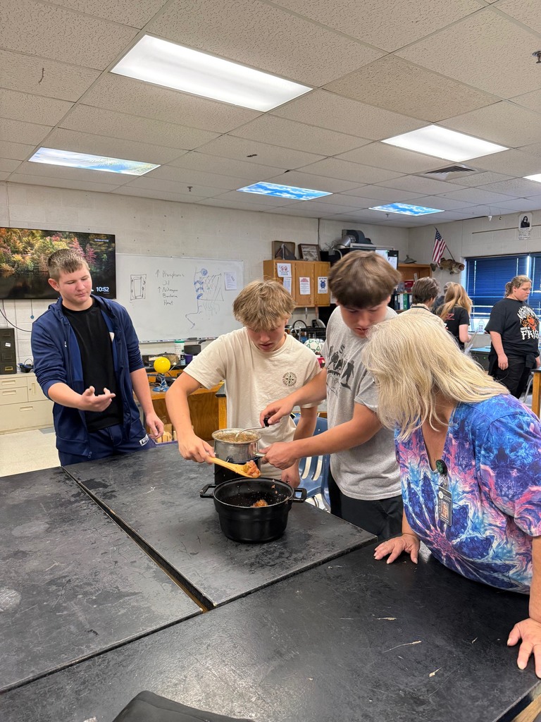 Students work together in a classroom, stirring apples in a pot as a teacher watches.
