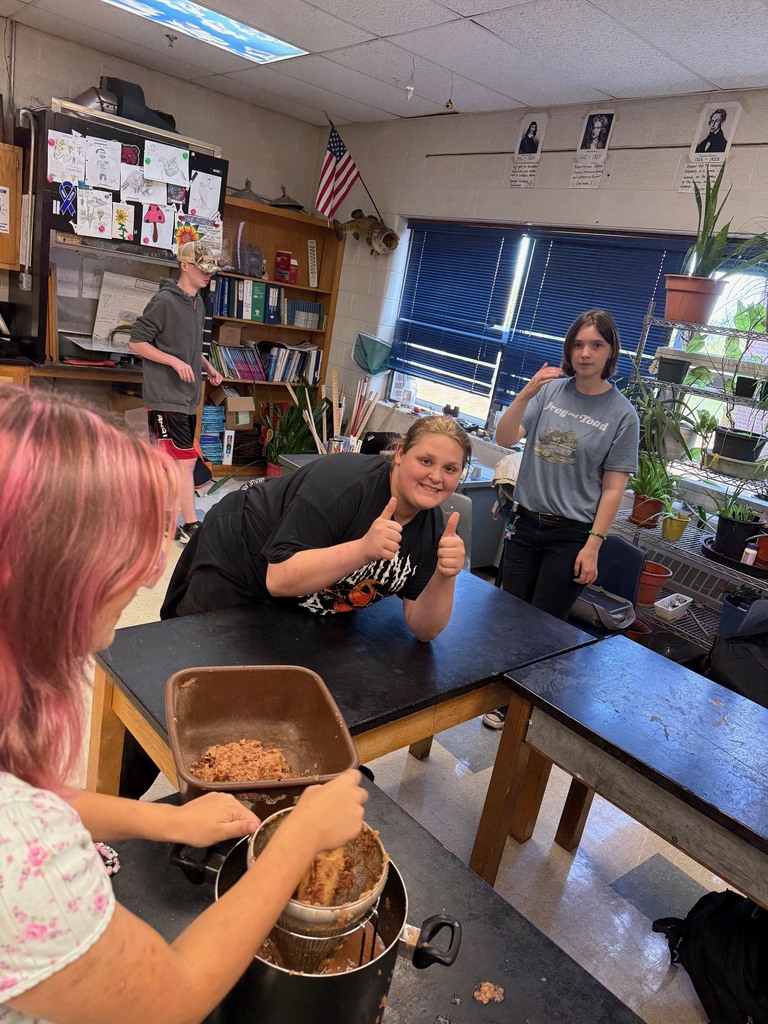 A student smiles and gives a thumbs up while classmates press apples through a strainer.