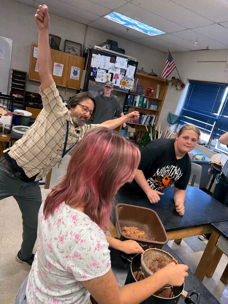 A teacher raises arms in excitement as students press apples through a strainer.