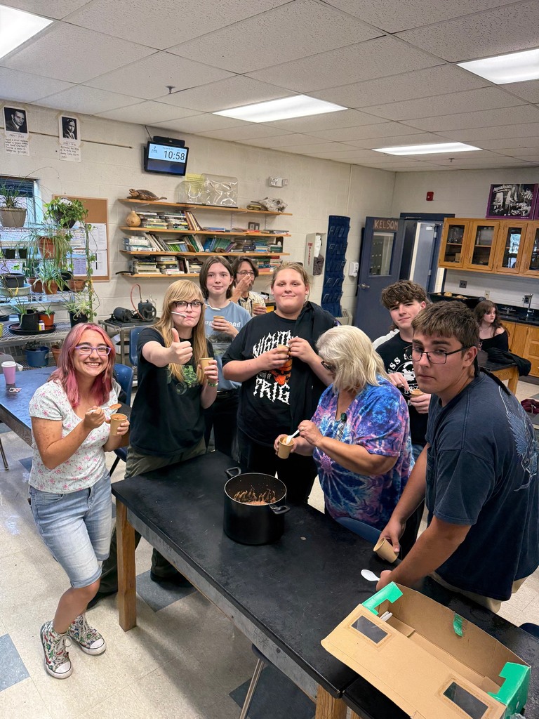 Students and a teacher gather around a table smiling and giving thumbs up while eating applesauce from cups.