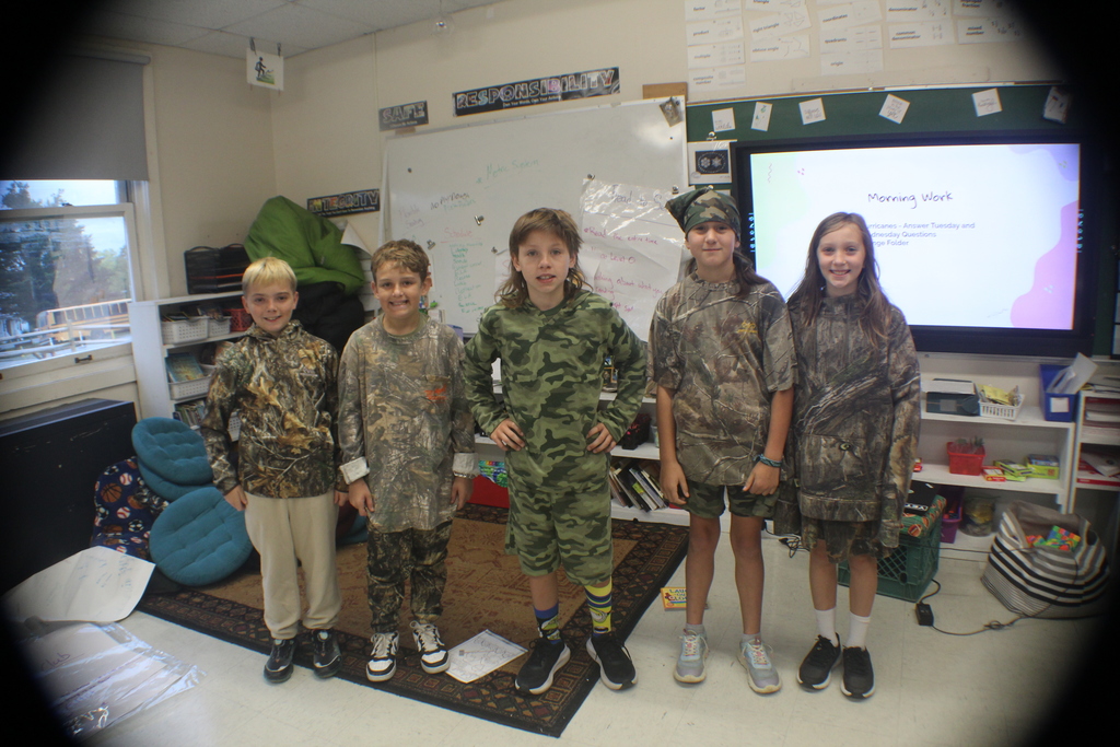 A group of five children wearing camouflage clothing stand together in a classroom.