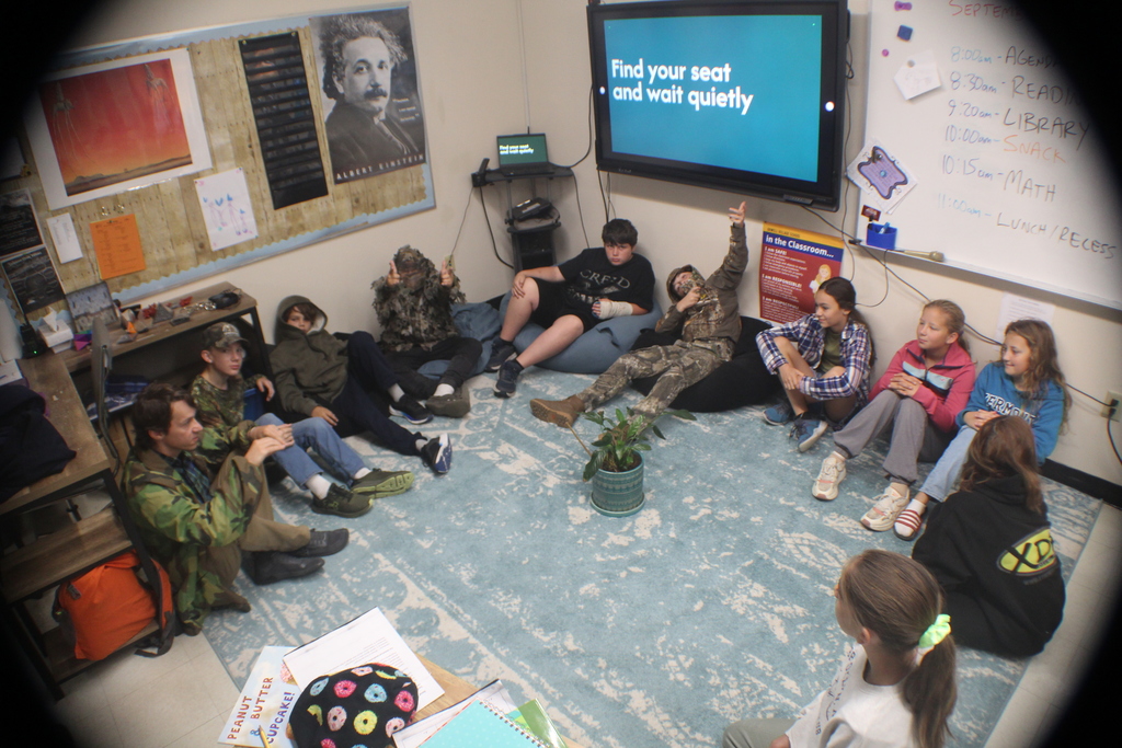 A teacher and a group of students are gathered on the floor in a classroom.