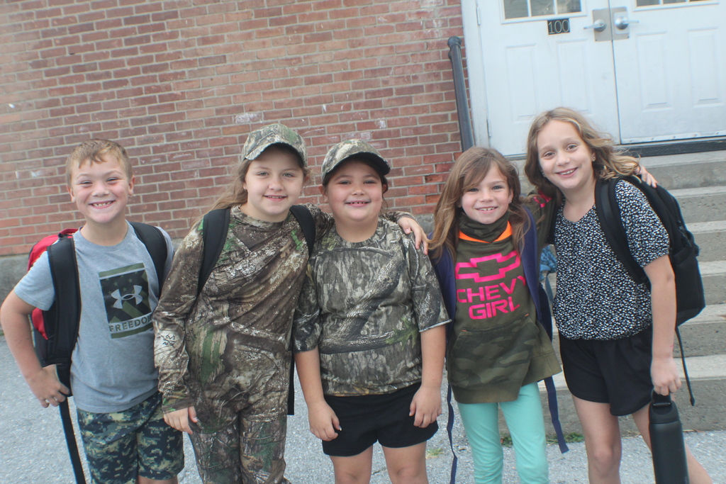 A group of five smiling children stands in front of a brick building.