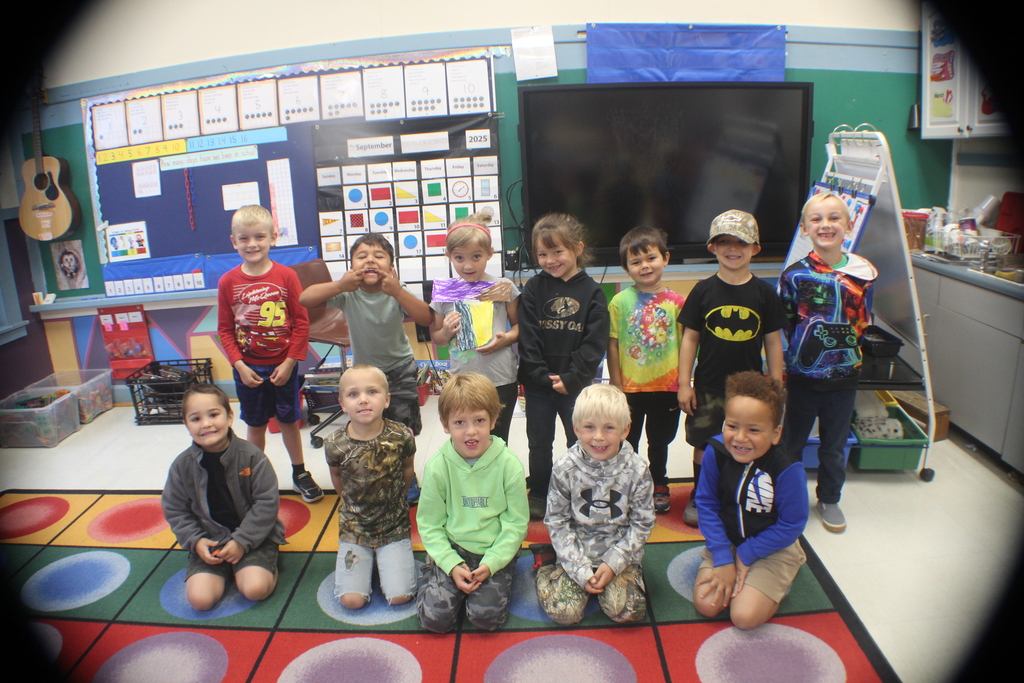 A group of nine smiling children, some standing and some kneeling, pose for a photo in a classroom.
