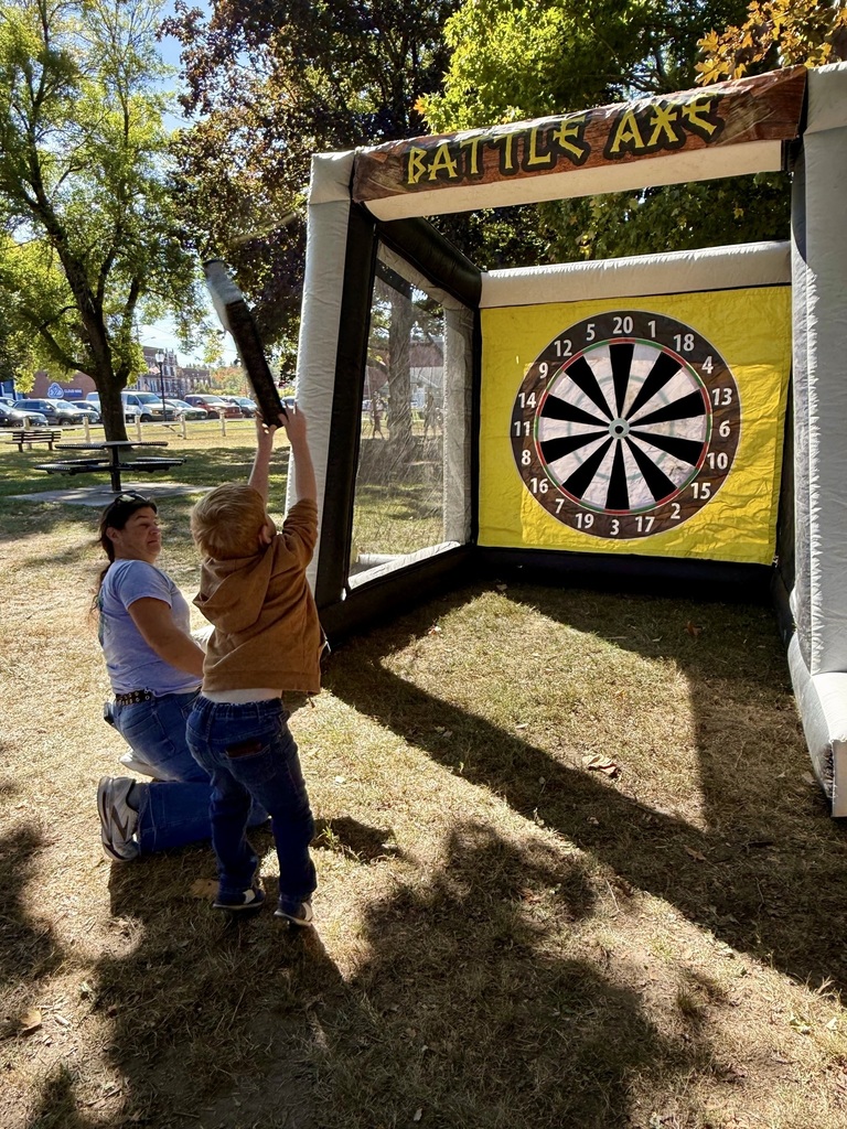 A young child throws a foam axe at a large inflatable target labeled “Battle Axe” while an adult kneels nearby for guidance.