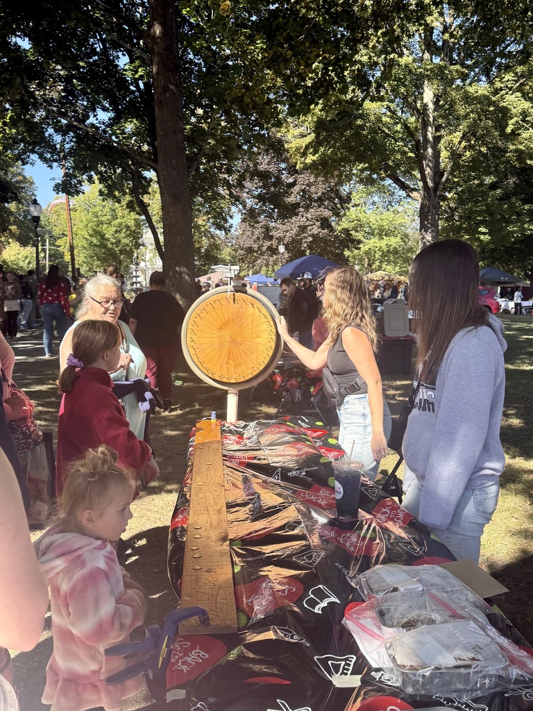 A crowd gathers at a booth where two young women run a spinning prize wheel game on a sunny day.