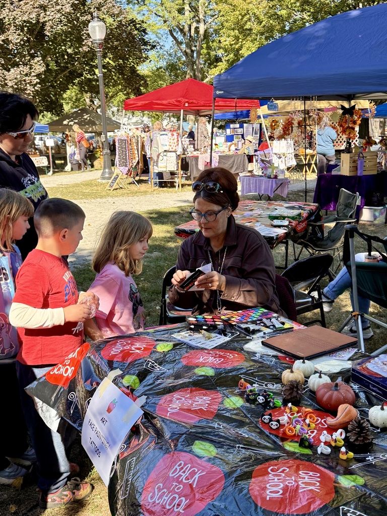A woman sits at a decorated table doing face painting for three children as another adult looks on at an outdoor festival.