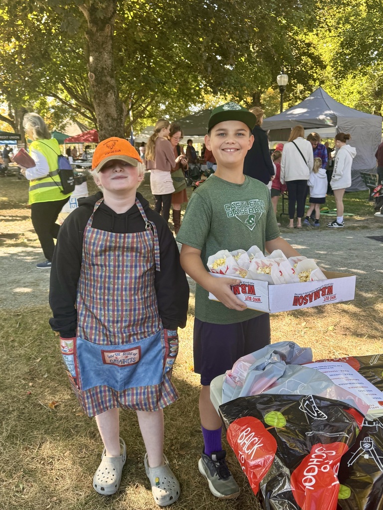 Two boys smile while selling popcorn; one wears an apron and orange cap, the other holds a tray filled with bags of popcorn.
