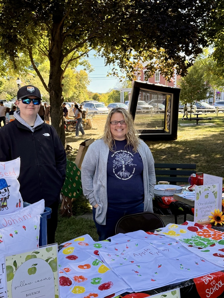 A man and woman stand behind a table covered with pillowcases painted with apple prints, sold as student artwork at a fair.