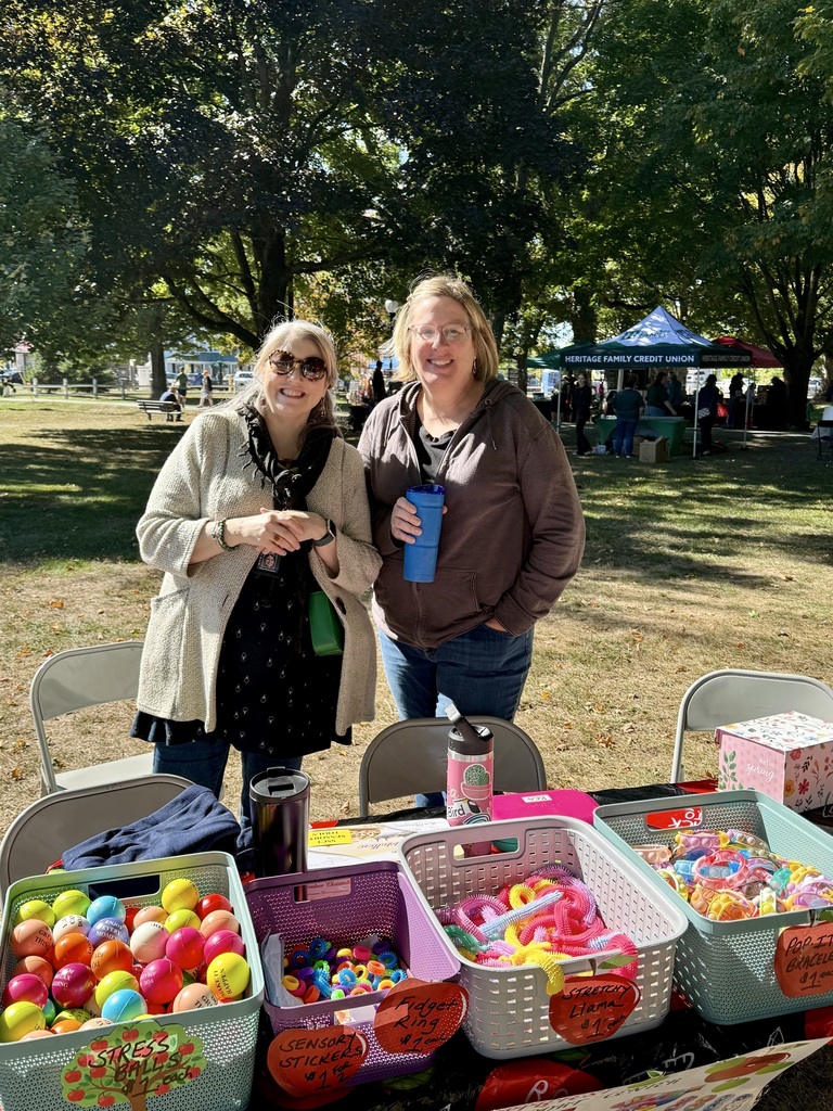 Two women smile at an outdoor booth with baskets of colorful toys labeled stress balls, sensory stickers, fidget rings, and stretchy llamas.