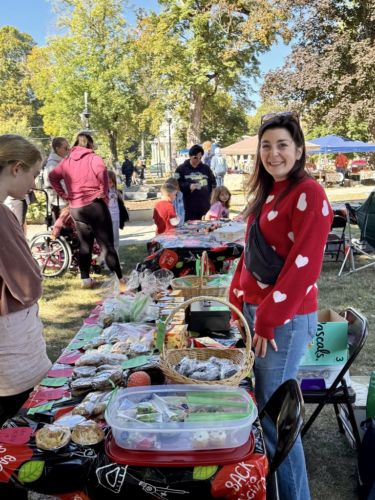 A woman in a red sweater with white hearts stands behind a table of baked goods wrapped for sale at an outdoor event.
