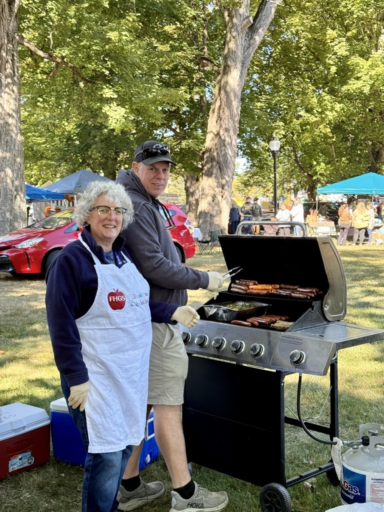 A man and woman grill hot dogs and sausages at a large barbecue grill during an outdoor community festival.