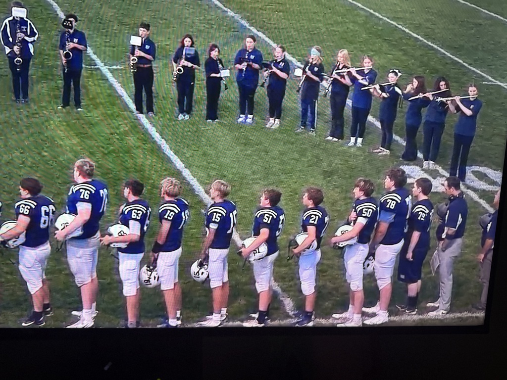 A row of football players in navy uniforms stand on the sideline holding helmets during the national anthem, with the school band lined up behind them playing instruments on the field.