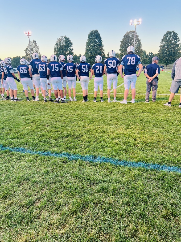 A line of football players in navy uniforms and helmets stand on the sideline of a lit field at dusk, facing the field.