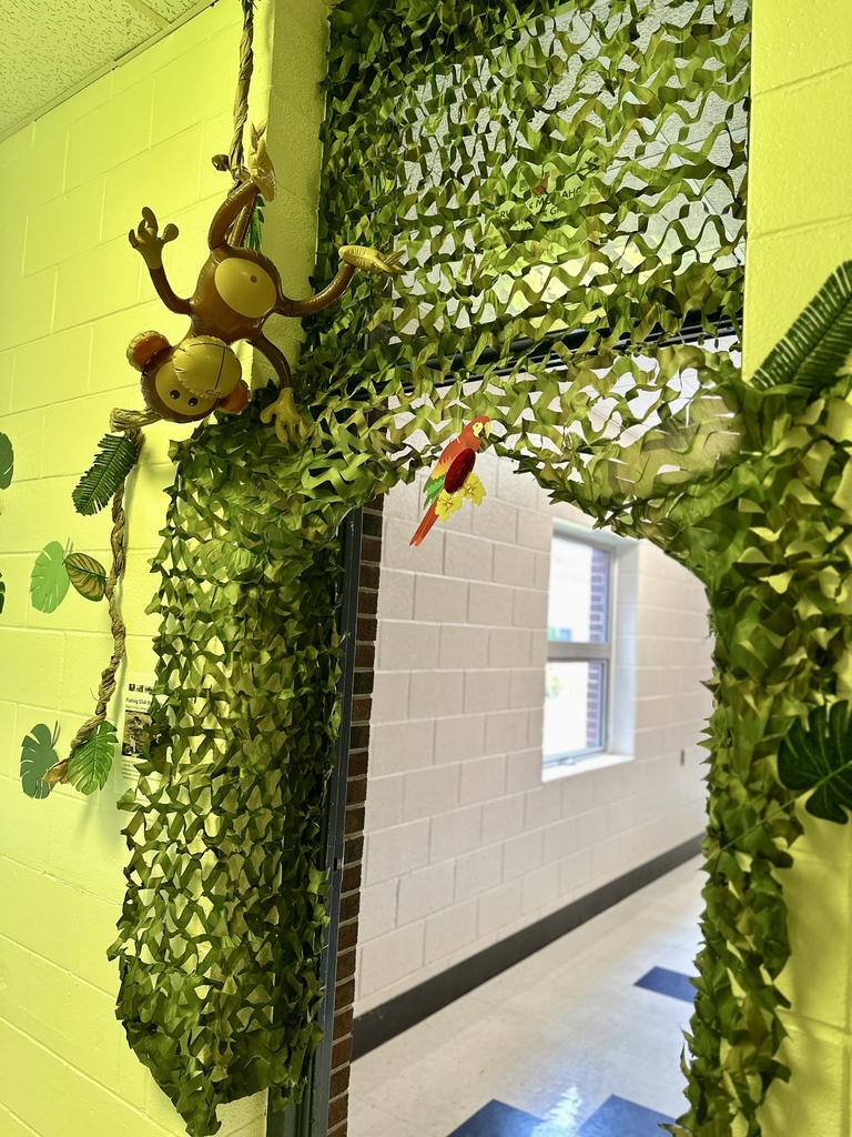 A classroom doorway framed in green netting with leaves and vines. A toy monkey hangs upside down from a rope, with a red parrot decoration nearby.