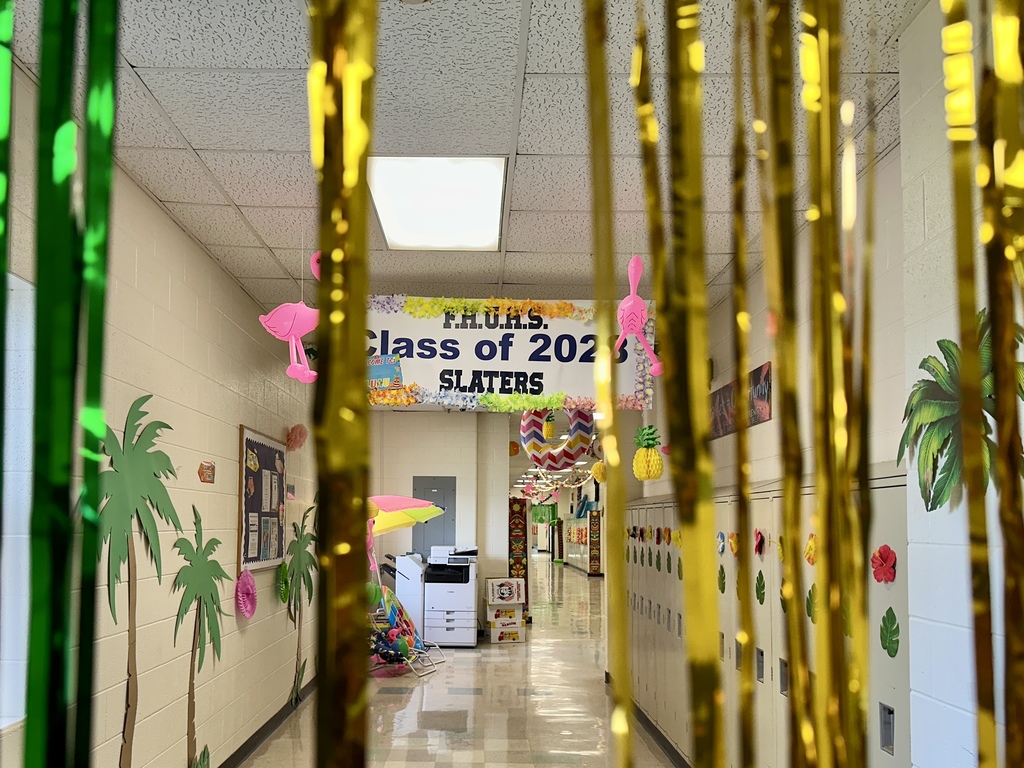 School hallway decorated with palm tree wall cutouts, pink flamingos, leis, pineapples, and a banner reading “FHUHS Class of 2026 Slaters.