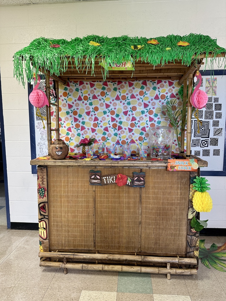 A bamboo tiki snack shack with a grass roof, colorful fruit-patterned backdrop, and tropical decorations.