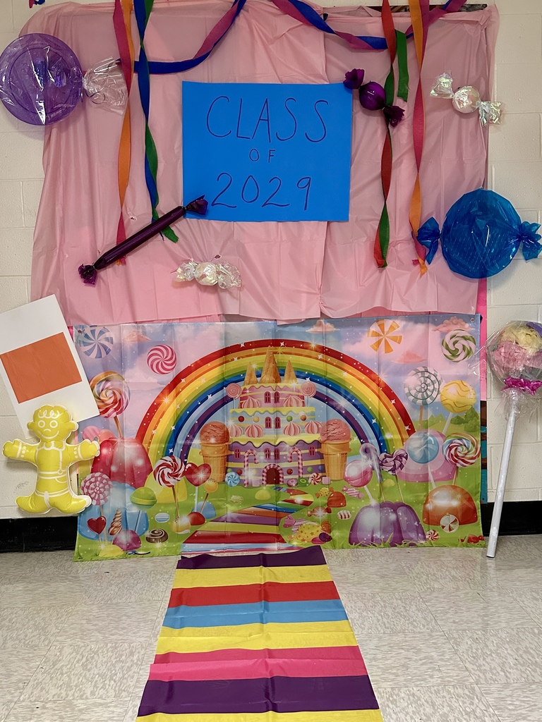A hallway display with a pink backdrop, rainbow carpet, large candy decorations, and a sign reading “Class of 2029.