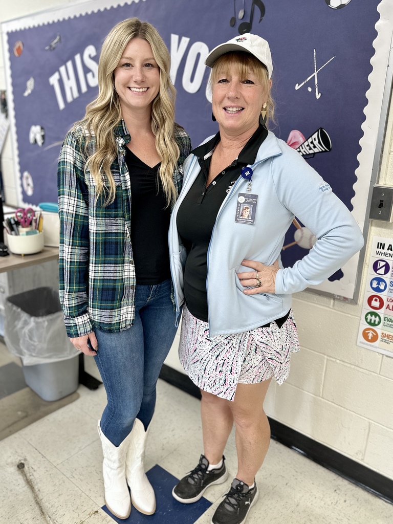 Two women posing together, one in a plaid shirt and jeans with white boots, the other in a light jacket, tennis skirt, and cap.