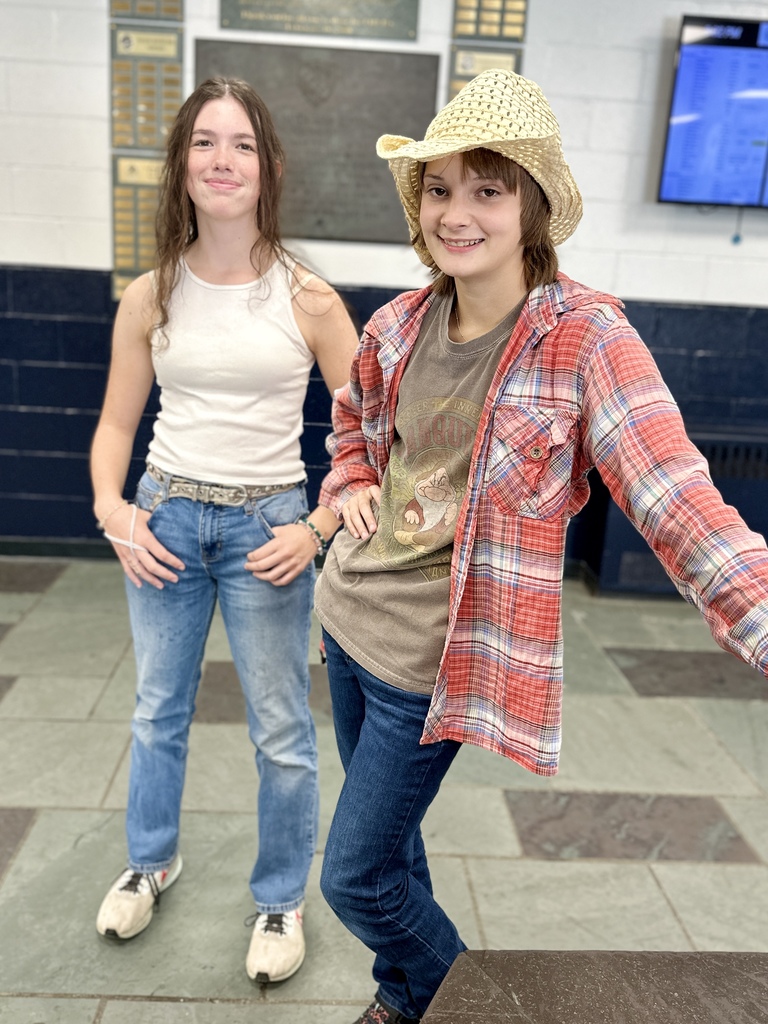 Two students standing together, one in a white tank top and jeans, the other in a plaid shirt, graphic tee, jeans, and cowboy hat.