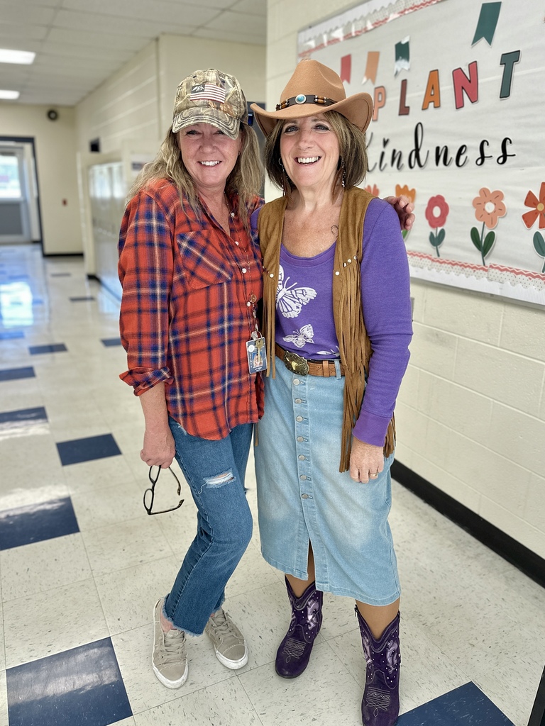 Two women dressed for spirit day, one in a plaid shirt and camo hat, the other in a cowboy hat, denim skirt, and purple boots.